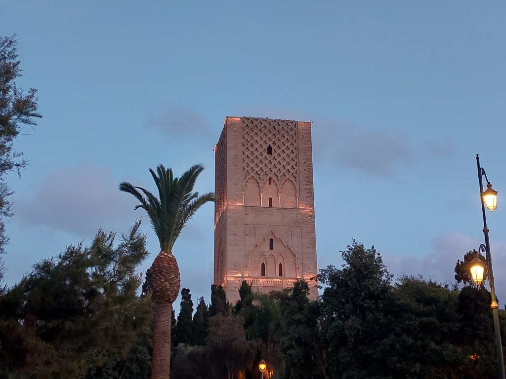 Hassan Tower in Rabat at sunset, lit up against a soft blue sky with palm trees and lanterns