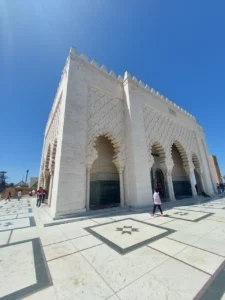 The white marble Mausoleum of Mohammed V in Rabat, Morocco, under a bright blue sunny sky, showcasing traditional Moroccan architecture and tourists walking on the esplanade.