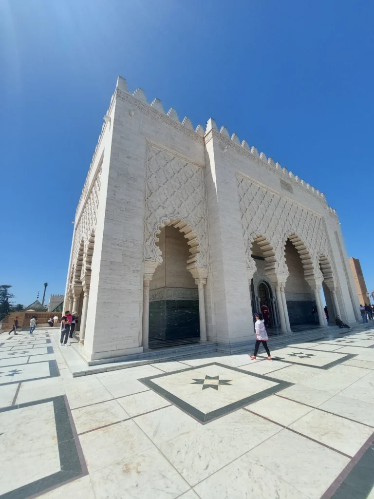 The white marble Mausoleum of Mohammed V in Rabat, Morocco, under a bright blue sunny sky, showcasing traditional Moroccan architecture and tourists walking on the esplanade.