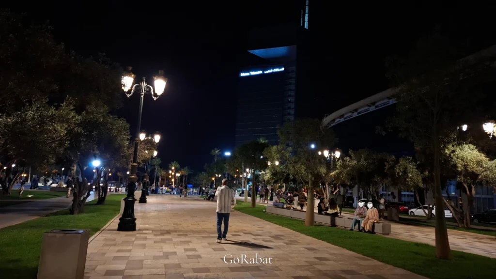A modern, well-lit pedestrian boulevard at night in Mahaj Riad, Hay Riad district of Rabat, Morocco, with people walking in summer clothing and the illuminated Maroc Telecom skyscraper.