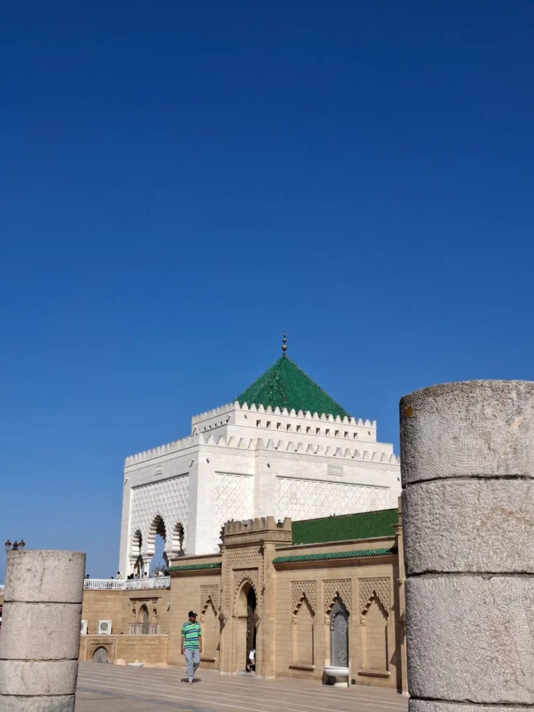 The white marble Mausoleum of Mohammed V with its iconic green tiled roof framed by ancient stone pillars under a clear blue sky in Rabat, Morocco.